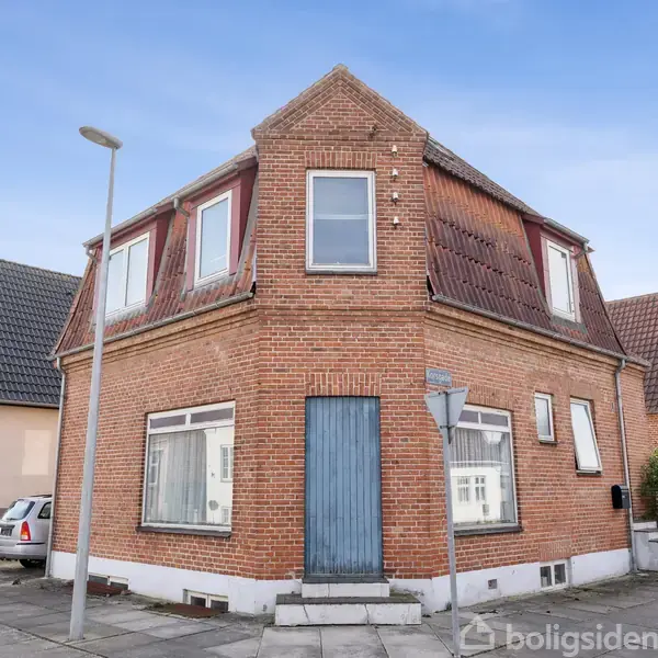 En to-etagers murstensbygning med en blå dør og røde vinduer står på et gadehjørne omgivet af andre huse. En lygtepæl ses i nærheden.