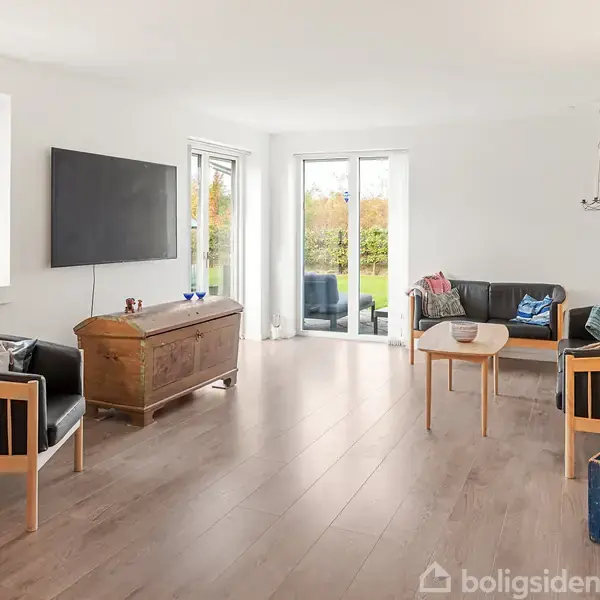 Stue med træmøbler og fladskærms-TV på væggen, sofa og stole omkring et sofabord, og store vinduer med udsigt til en grøn have. En blå kasse står på gulvet.