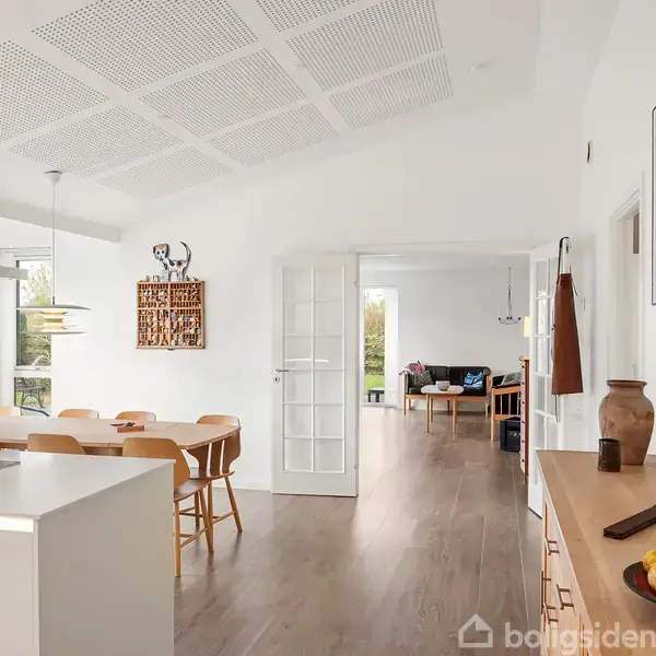 Køkkenalrum med træbord og seks stole står i et lyst rum med parketgulv. I baggrunden ses en stue med sofaer. En kandelaber og kunstværker dekorerer væggene.