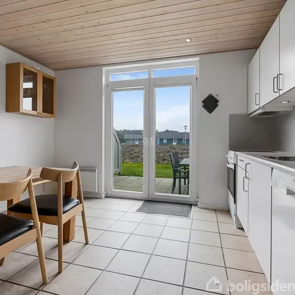 Køkken med hvidt interiør står stille. Et spisebord med tre stole er til venstre. Store glasdøre fører ud til en have. Vægur og lampe hænger på væggen.