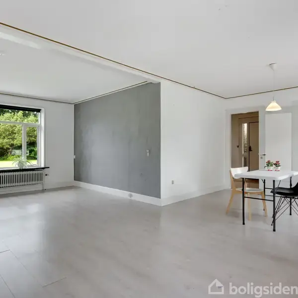 Et lyst rum med en grå væg og stort vindue mod venstre. En rund hvid bord står med to stole foran en åben dør til et andet rum.