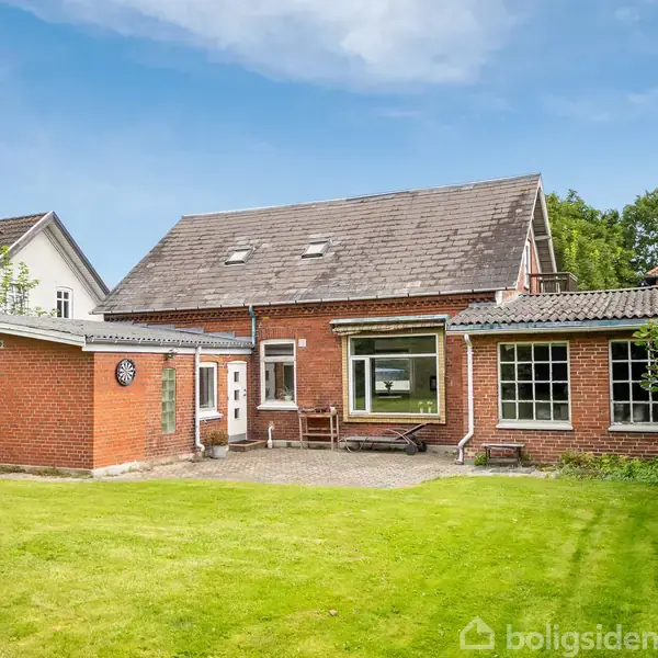 Rødstenshus med skrånende tag står stille i en grøn have. Stor vinduesrude og mindre sidebygning. Græsplæne omkranser, og himlen er blå.