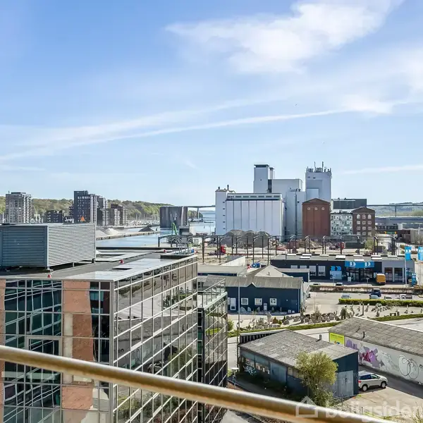 Udsigt over en by fra en balkon, hvor moderne bygninger og industriområder dominerer landskabet. Havnefronten kan ses i baggrunden med siloer og kajanlæg.