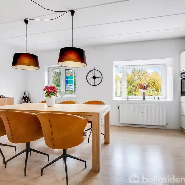 Et spisebord med fire brune stole står centralt i et lyst køkken. Over bordet hænger to lamper. I baggrunden er der et vindue med udsyn til grønne træer.