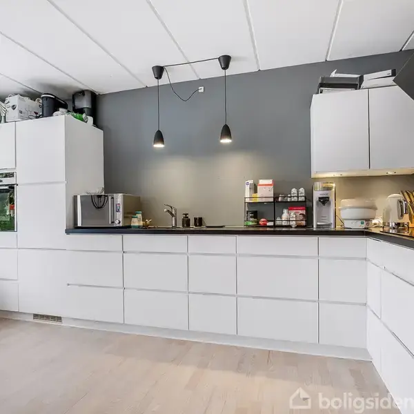 Køkken i moderne stil med hvide skabe og skuffer. Indbygget ovn, mikroovn og kaffemaskine på bordpladen. To pendellamper hænger over arbejdsfladen. Lyst trægulv og grå væg som baggrund.