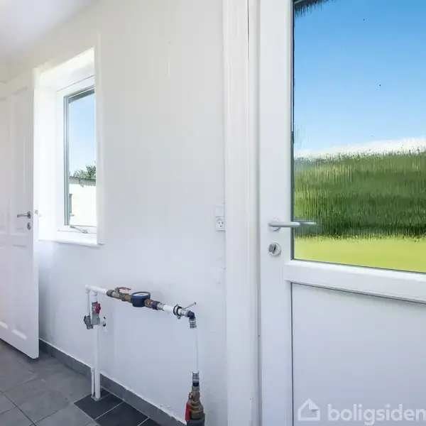 Dør åbner ud til en grøn græsplæne gennem et frostet glas. Indendørs ses hvide vægge, et flisegulv og en åben dør mod et lyst køkken. Vandrør monteret på væggen.