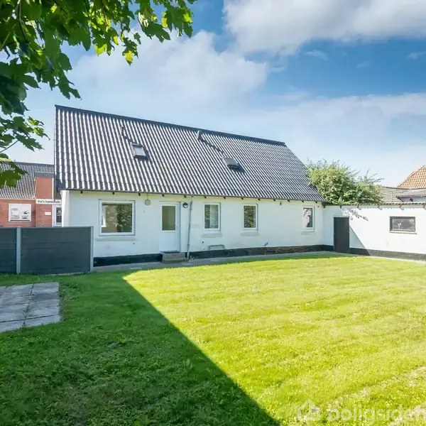 Hvidt hus med sort tag står stille omgivet af en velplejet græsplæne og nogle fliser i en solrig have; træer og hegn er i baggrunden.