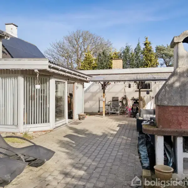 To liggestole står på en terrasse ved et hus med store vinduer. I baggrunden ses en grill i mursten og træer, hvilket skaber en hyggelig udendørs atmosfære.