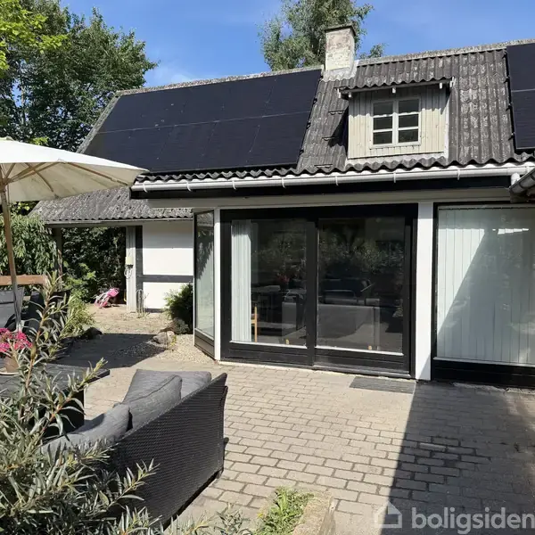 Et moderne hus med solpaneler på taget og store glasdøre. Foran huset er en terrasse med sofaer og en parasol. Grønne træer omkranser gårdspladsen under en klar blå himmel.