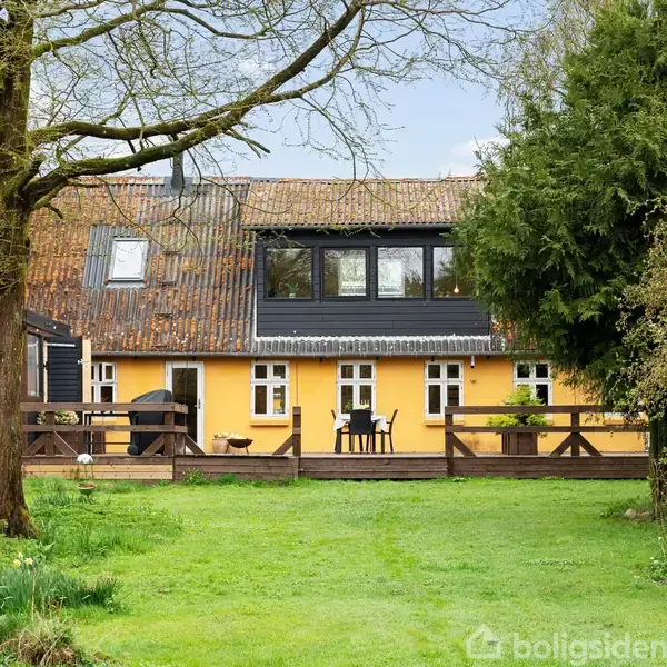 Gulmalet hus med sort tag i to etager. Husets terrasse har møbler og en grill. Omkring huset er der frodig have med græsplæne, træer og buske.