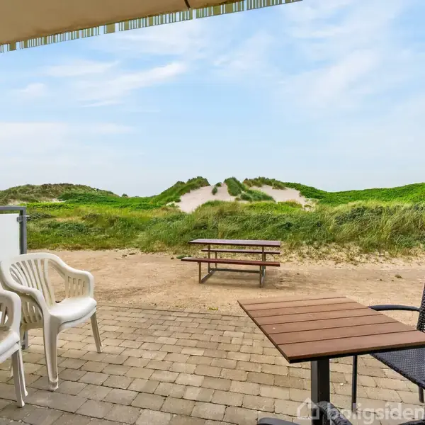 Terrassemøbler står på en flisebelagt terrasse. Plaststole og et træbord vender mod en sandklit med græs. En bænk står i sandet i baggrunden under en blå himmel.