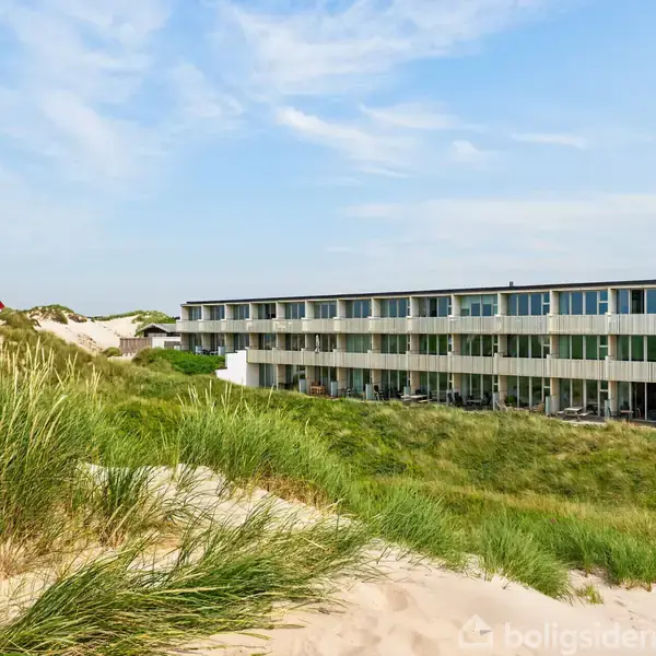 En lang, moderne bygning står i sandklitterne, flankeret af tæt græs. Til venstre ses et rødt, trekantet skulptur. Blå himmel over sand og græs skaber en naturlig atmosfære.