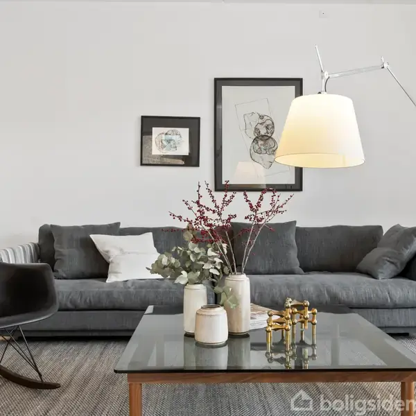 Grå sofa står mod en hvid væg med to indrammede billeder. Sort gyngestol og hvid stol flankerer et glasbord med planter og dekorationer. Hvid stålampe til højre.