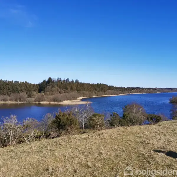En flod strømmer stille gennem et landskab med skovklædte bakker og åbne marker under en klar blå himmel.