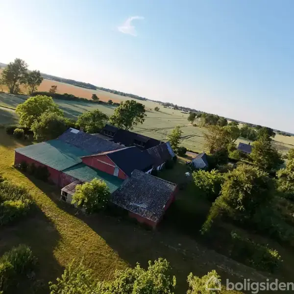 En gård ligger midt i åbne marker og omgives af grønne træer, med små bygninger spredt omkring. Marken strækker sig til horisonten under en klar blå himmel.