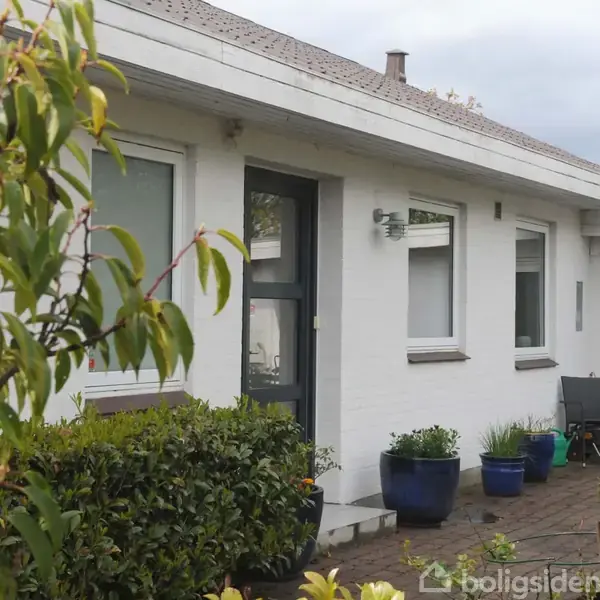 Terrassemøbler med parasol står på flisebelagt område foran hvidmalet husvæg med vinduer. Omkring er blå potteplanter og en hækkant. Lysindfald foregår under overskyet himmel.