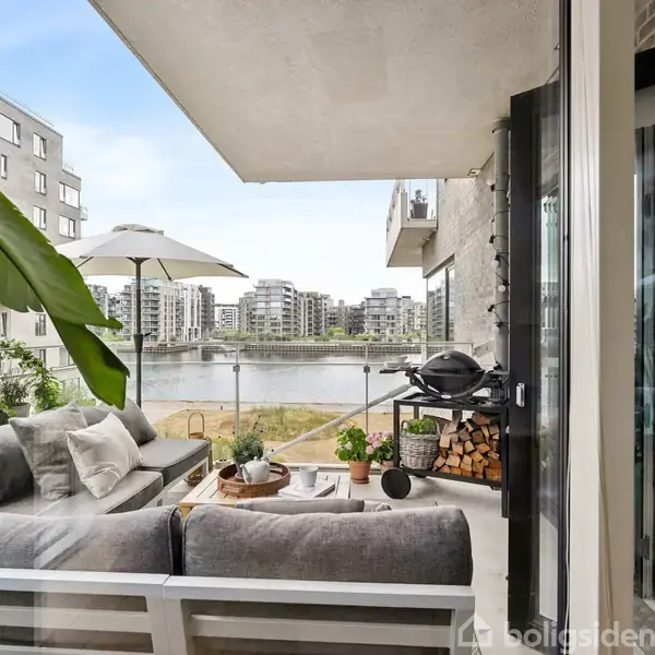 Balkon med sofa og bord, placeret med udsigt over en fredelig flodbyen. Omgivet af moderne lejlighedsbygninger og udstyret med gasgrill og parasol til afslapning.