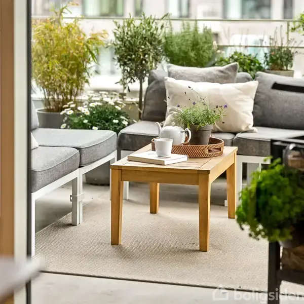 En træbord står på en altan omgivet af grå sofaer med puder. På bordet er en bakke med en plante og en kop. Baggrunden har grønne planter.