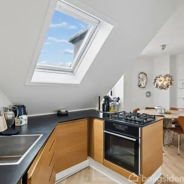 Et køkken med skråt loftsvindue viser sort køkkenbord med vask, komfur og kaffemaskine. I baggrunden ses et spisebord med brune stole og en dekorativ lampe.