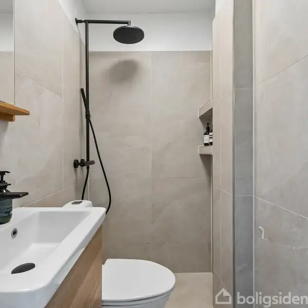 En moderne bruser hænger på en vægflise i et lille badeværelse. Foran står en hvid vask med en sort sæbedispenser, omgivet af lyse beige fliser.