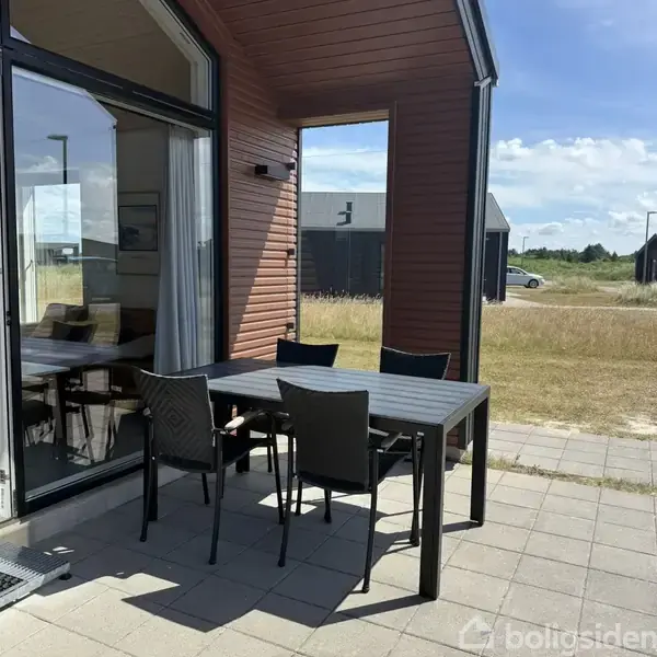 Et bord med fire stole står på en terrasse. Bordet er tomt, og terrassen er ved et moderne træhus med store vinduer. I baggrunden ses en græsplæne og en parkeringsplads.