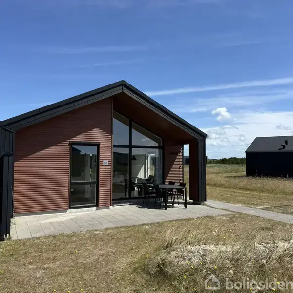 Et moderne sommerhus med store vinduer står på en flisebelagt terrasse. Det er omgivet af en græsplæne under en blå himmel med skyer.