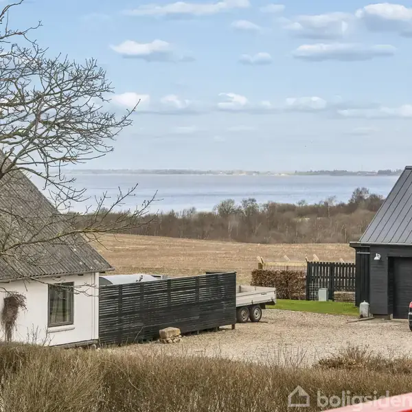 Et hus med sort tag og en trailer står på en gårdsplads ved siden af en sort bil. Omkringliggende marker og en sø ses i baggrunden under en blå himmel.