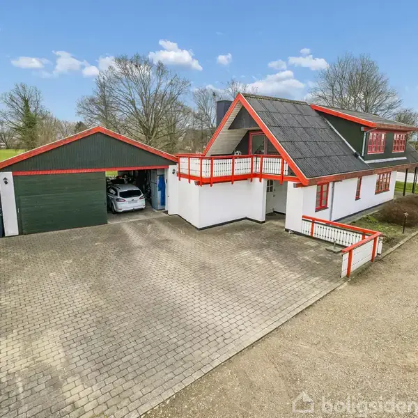 Der er et hvidt hus med rødtag og balkon. Ved siden af er en grøn garage med en bil. Omgivet af flisebelagt gårdsplads og et landligt landskab.