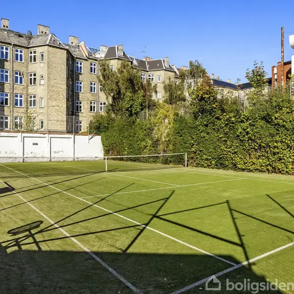 En grøn tennisbane står ubenyttet i solskin omgivet af høje bygninger med mange vinduer og tætte grønne buske på siderne. Skrevet tekst: "boligsiden".