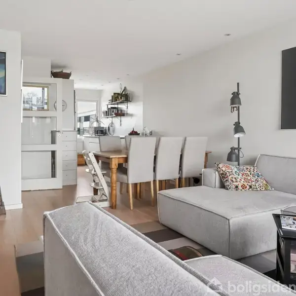 Stue med en grå hjørnesofa og sofabord, lamper ved siden. I baggrunden ses spisebord med stole og væghylder. Væggene er pyntet med kunstværker. Gulvet er lyst træ.