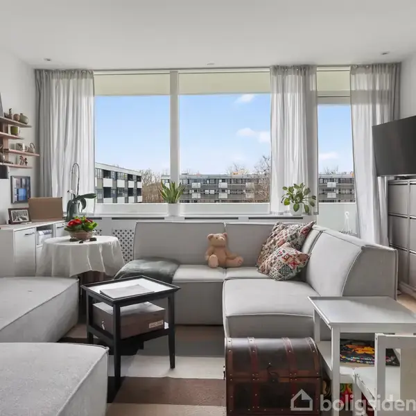 Stue med lysegrå sofa i vinkel fyldt med farverige puder, stående foran stort vindue med hvide gardiner. Rummets dekoration inkluderer planter, træmøbler og en væghængt reol.