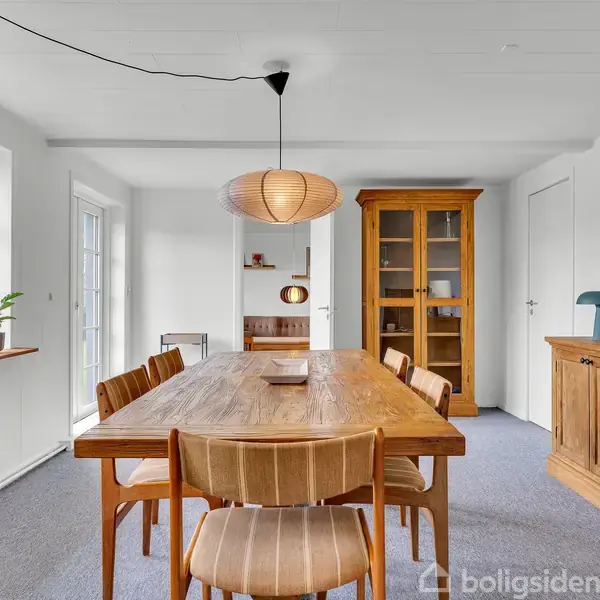 Et rektangulært spisebord med seks stole står i en lys spisestue. En loftslampe hænger over bordet. I baggrunden er et skab, en vægkonsol og vinduer, som tillader dagslys.