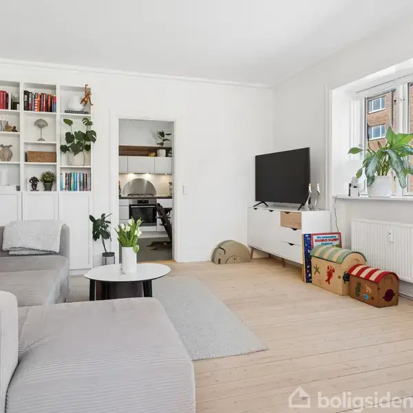 Stue med lys sofa, reol fyldt med bøger og planter, samt et fladskærms-tv på et mediemøbel. Rummet har trægulv, planter ved vinduet og adgang til et køkken.