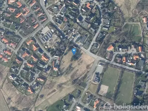 Byområde med huse set oppefra, placeret langs veje, omgivet af marker og grønne arealer; en blå prik markerer en placering midt i billedet.