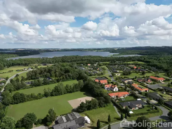 Bebyggelse med huse omgivet af grønne marker strækker sig mod en stor sø i baggrunden. Skyer dækker delvist himlen, hvilket skaber en fredfyldt, landlig atmosfære.