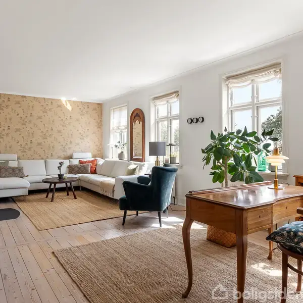 En lys stue med træmøbler og planter. En stor, hvid sofa står ved væggen, mens et skrivebord med stol er ved vinduerne. Et tæppe dækker trægulvet.