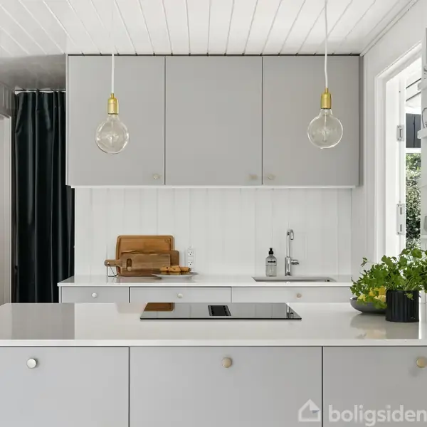En moderne køkkenø med en indbygget kogeplade står i et lyst, minimalistisk køkken. Over hænger to pærer. Skærebrætter og en urtepotte pynter på bordpladen.