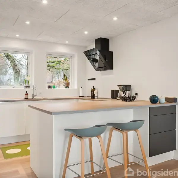 Et moderne køkken med køkkenø, to barstole og hvid indretning. Kogeplader og emhætte er centrale. To vinduer giver udsigt til træer udenfor. Ingen tekst findes.