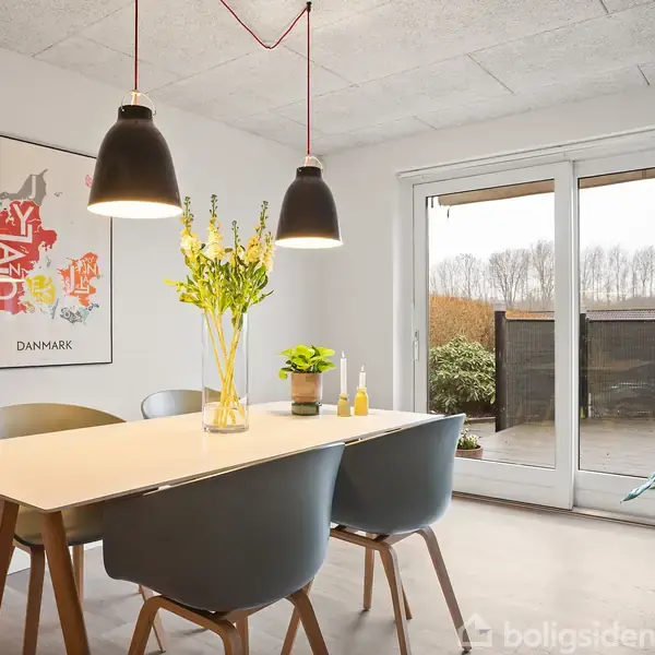 Et spisebord med fire stole er centreret i et lyst rum. To pendellamper hænger over bordet, som har blomster og lysestager. Til venstre hænger et Danmark-billede.