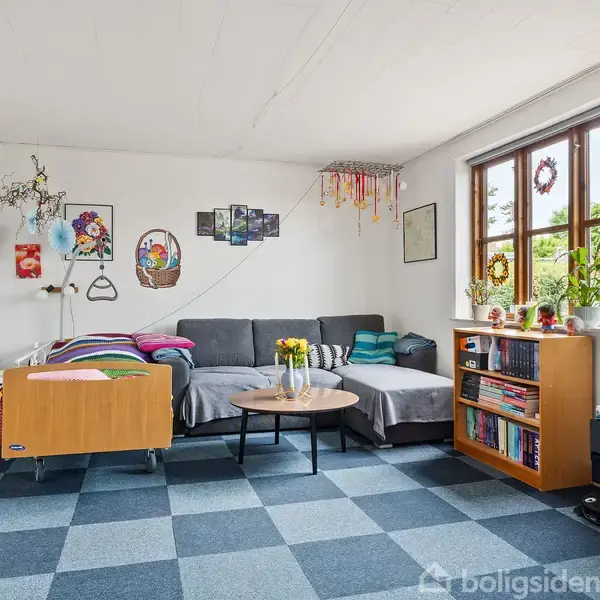 Stue med grå hjørnesofa og rundt sofabord, pyntet med blomster. Væggene har dekorative billeder, og der står en bogreol ved vinduet med udsigt til have. En farverig plaid ligger på sofaen.