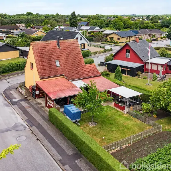 Gult hus med rødt tegltag står på hjørnegrund. Grøn have med træer og skure omgiver det. Nabohuse i varierende farver er synlige i et roligt boligområde.