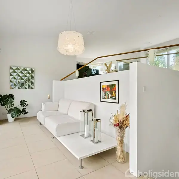 Hvid sofa står i moderne stue med et vægmaleri over sig. Store vinduer kaster lys på flisegulve. Rundt om er grønne planter, vægkunst og en indbygget pejs med glasdør.