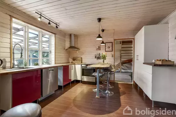 Køkken med hvide trævægge og røde skabe. En køkkenø med barstole står midt i rummet. Lysindfald fra vinduer, træloft med lamper, og hvidt skab i hjørnet.