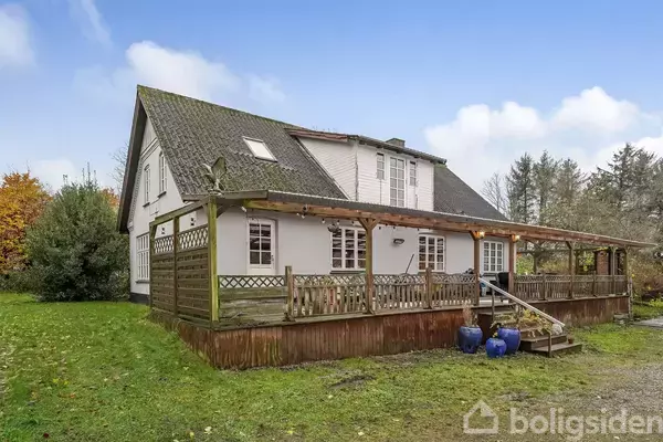 Et hus med træterrasse og veranda. Terrassehuset er omgivet af en grøn have med buske og træer, og himlen er klar og blå. Tre blå krukker står ved trappen.