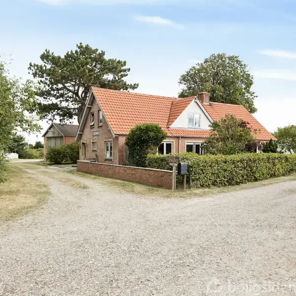 Et rødt murstenshus med rødt tegltag står omgivet af hække og træer. Det ligger ved en grusvej i et landligt område med blå himmel.