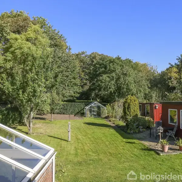 Fladskærms-TV står på reol, viser ingen billede; flere hylder har pynt. Stuen har spisebord- og stole, vægkunst og store vinduer, der giver naturligt lys.
