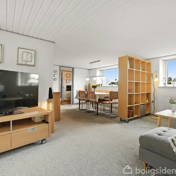 En glasoverdækket stue med en blomsterprintet sofa og et træbord. Bagved ses en have med grønne træer. En dør til højre åbner ud til terrassen.
