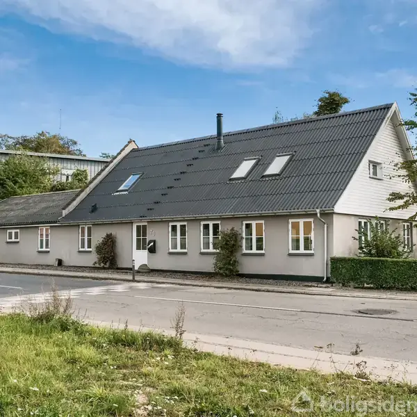 Et lang, beige hus med sorte tagsten står langs en landevej, omgivet af grønne træer og buske. Himlen er klar med spredte skyer.