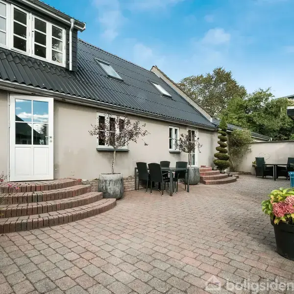 Et hus med en mørkt tag og lyse vægge er omkranset af en flisebelagt terrasse. Der står havemøbler og planter i potter. Himlen er klar med nogle få skyer.