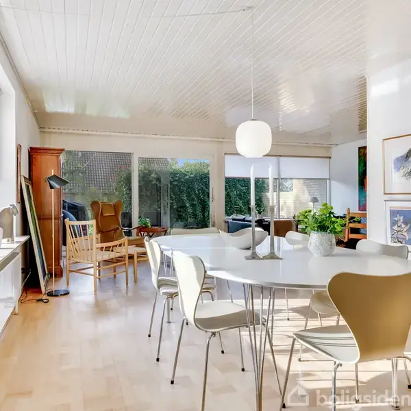 Et lyst spisebord med hvide stole står i en rummelig spisestue. Store vinduer giver udsyn til en grøn have. Væggen er dekoreret med kunstværker.